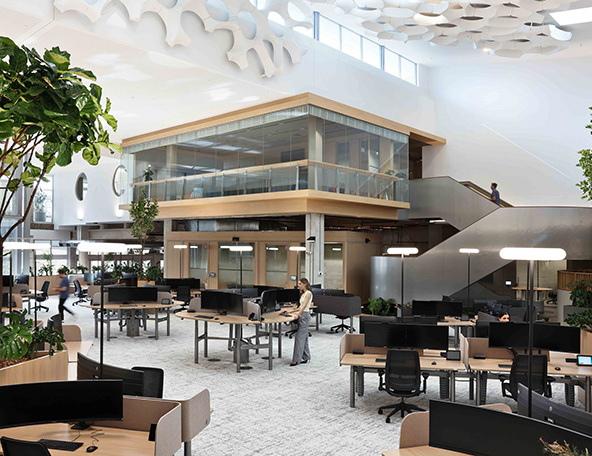 A modern office space featuring open work areas, multiple desks with computers, greenery, and a glass-walled upper level with stairs.