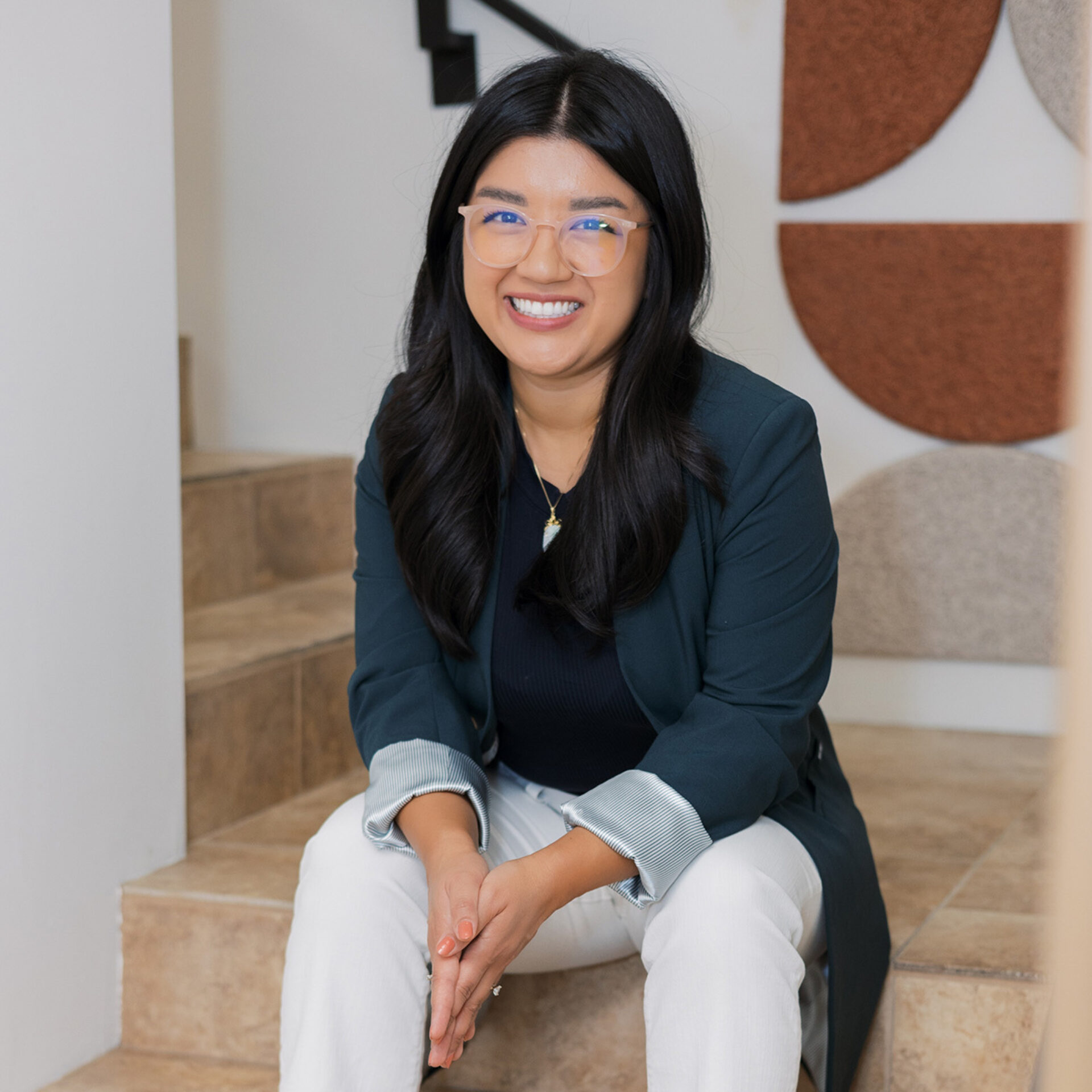 Smiling person with long dark hair and clear glasses wearing a teal blazer, black top and white pants, seated on tiled stairs, hands clasped.
