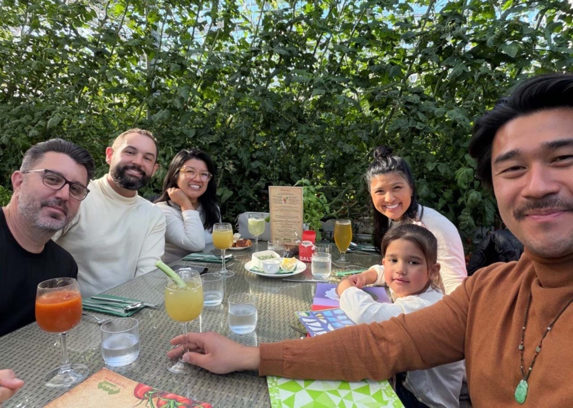 Six people—four adults and a young girl—smile at a sunlit patio table beneath dense green vines, surrounded by cocktails, glasses and menus.
