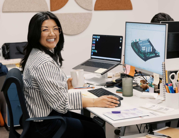Smiling woman in striped shirt at a cluttered desk holds a white mug while using a mouse; two monitors show a laptop and 3D site model.