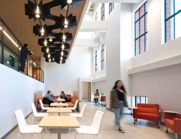 Bright two-story campus lounge with tall windows, black pendant lights, white tables, orange couches, students studying and walking.