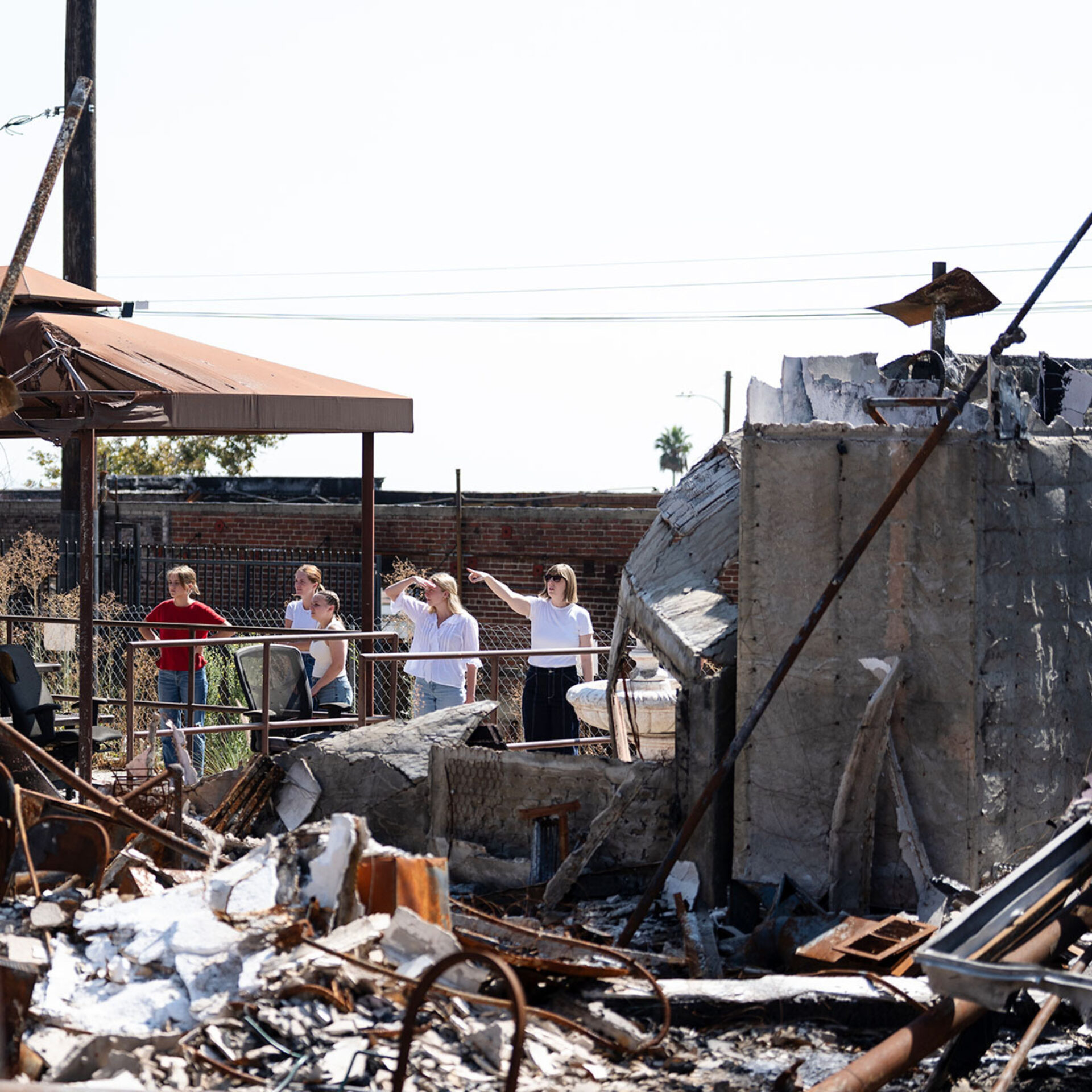 Five people on a raised walkway under a rusted canopy survey charred rubble, collapsed concrete walls and twisted metal after a fire.