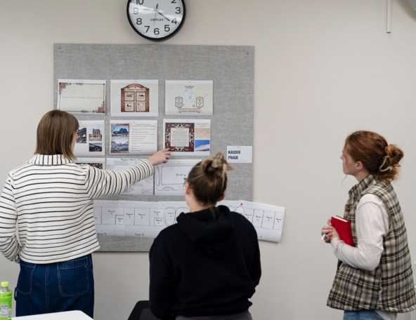Three women study pinned design layouts on a gray bulletin board under a wall clock; one in a striped sweater points at a printed plan.