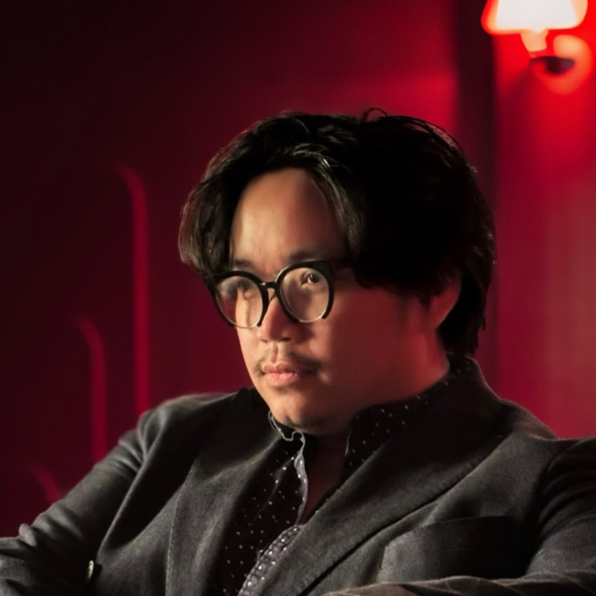 Seated man with round black glasses, wavy dark hair and faint mustache, wearing a gray blazer and dotted shirt under warm red lighting.