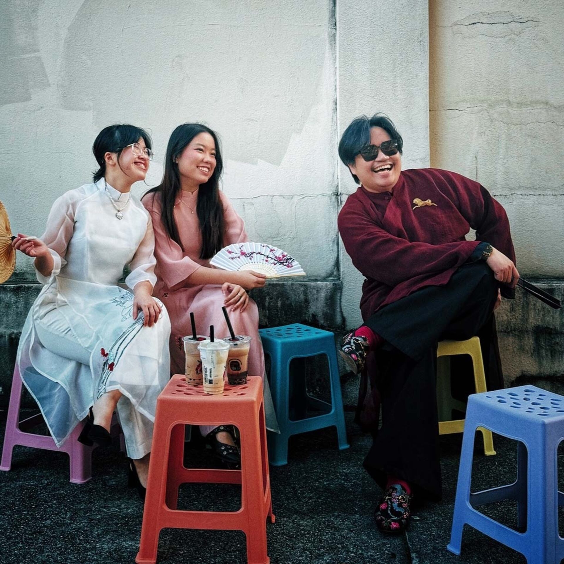 Three friends on plastic stools by a cracked wall; two women in long dresses with fans and boba, a laughing man in a maroon robe and shades.