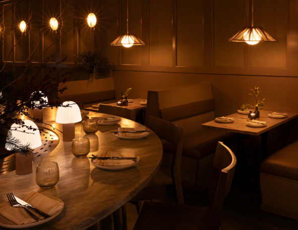 Warm, dim restaurant interior with a curved marble bar, glowing tabletop lamps, rattan pendant lights, booth seating and set place settings.