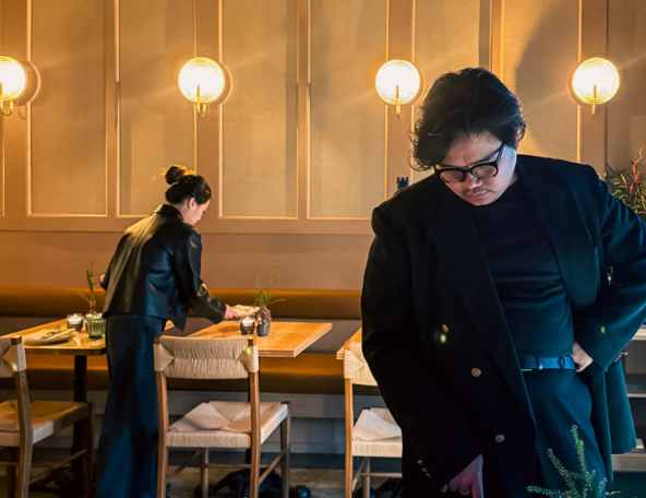 Warm-lit restaurant interior with a woman in a leather jacket arranging a table and a bespectacled man in a black blazer looking down.