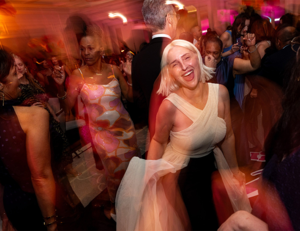 Crowded reception dance floor with a short blonde woman in a white sleeveless dress laughing, tulle swirling under warm colorful lights.