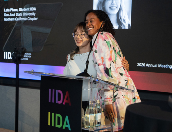 Two women smile and embrace at an IIDA podium; a woman with glasses stands beside a woman with curly hair wearing a colorful floral blazer.