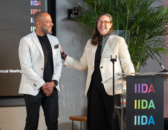 Two smiling presenters at a podium for an IIDA event: woman with glasses and beige blazer gestures toward a laughing man in a light blazer.
