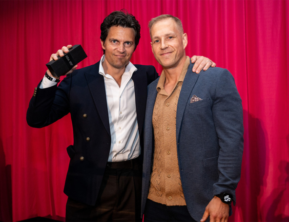 Two men in blazers stand before a pink curtain, one holding a black award box aloft while the other rests a hand on his shoulder, smiling.