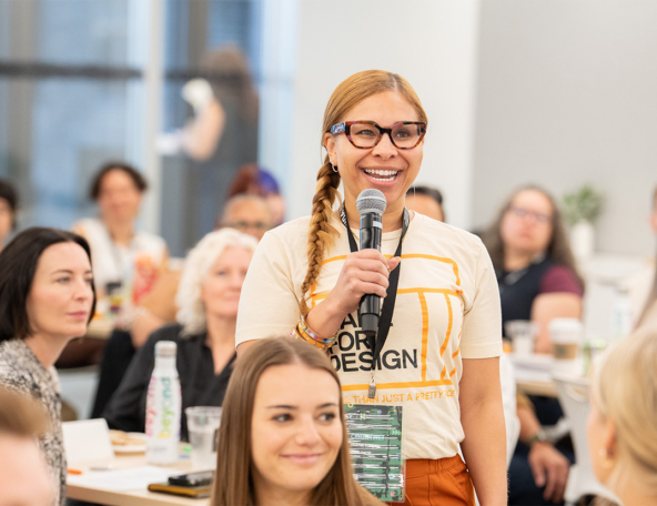 Smiling woman with braided hair and tortoiseshell glasses holds a microphone while speaking at a packed design workshop, lanyard visible.