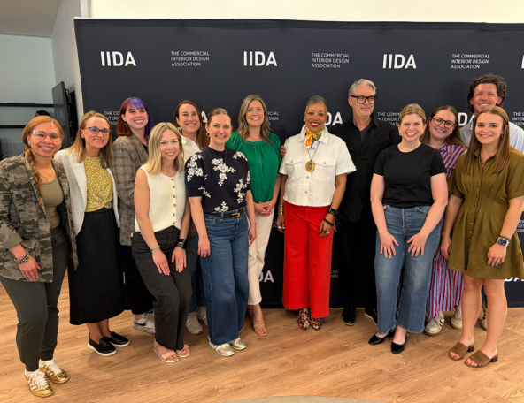 Diverse, smiling interior designers pose before a black IIDA backdrop; one woman wears bright red pants, white jacket and a medallion.
