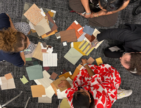 Overhead view of four people seated on gray carpet arranging assorted fabric, wood and tile swatches; one person wears a red floral dress.