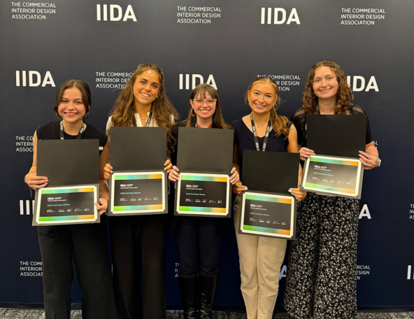 Five smiling young women stand shoulder-to-shoulder holding framed certificates and wearing lanyards against a navy IIDA awards backdrop.