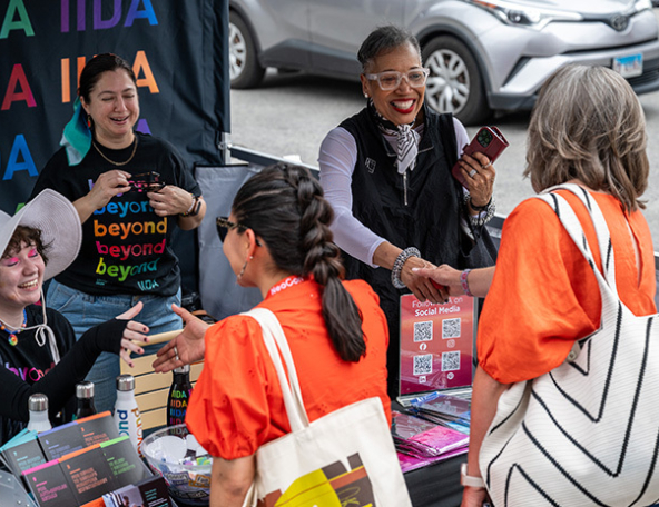 Smiling vendors behind a colorful IIDA booth greet two women in orange shirts, exchanging handshakes, tote bags and event swag.