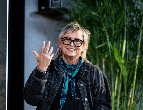 Smiling older woman with short blonde hair and black glasses, wearing a denim jacket and teal scarf, waving and showing rings.