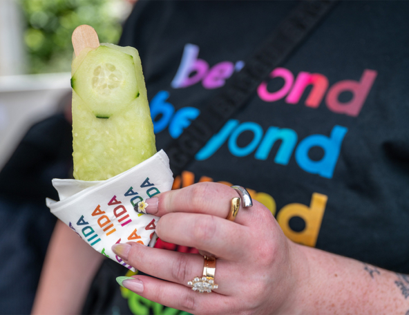 Hand with gold and silver rings and a daisy nail holding a pale green cucumber popsicle wrapped in multicolored branded paper.
