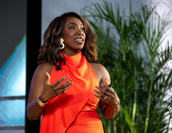 Woman in a sleeveless, bright orange dress gestures expressively while speaking on stage, wearing gold hoop earrings and a clip mic.