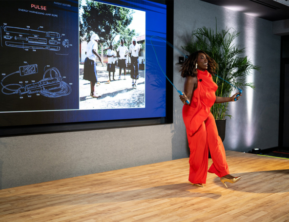 Woman in a bright red jumpsuit energetically jumping rope on a wooden stage beside a large projection showing a patent diagram and a photo.