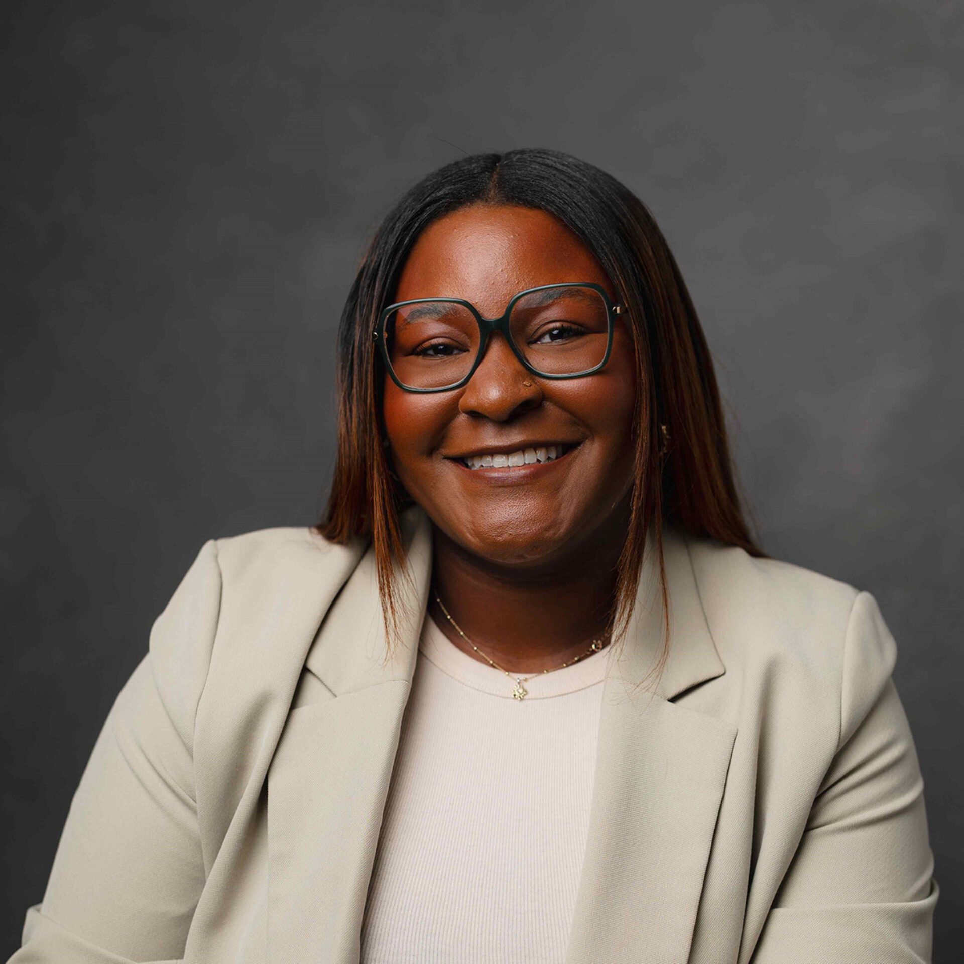 Smiling Black woman with straight shoulder-length hair, teal-rimmed glasses, cream blazer and small pendant necklace on a gray backdrop.