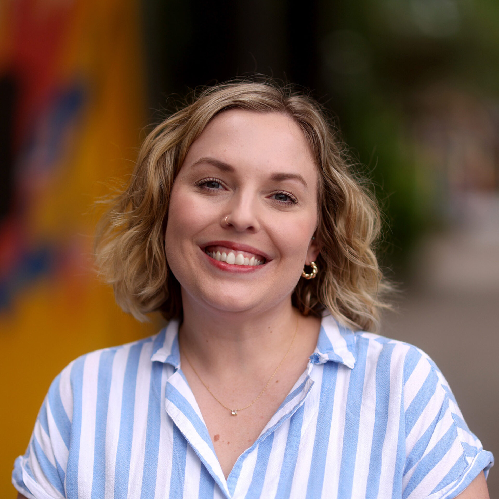 Woman with short wavy blonde hair and nose ring smiles warmly wearing a blue-and-white striped shirt and small gold hoop earrings.