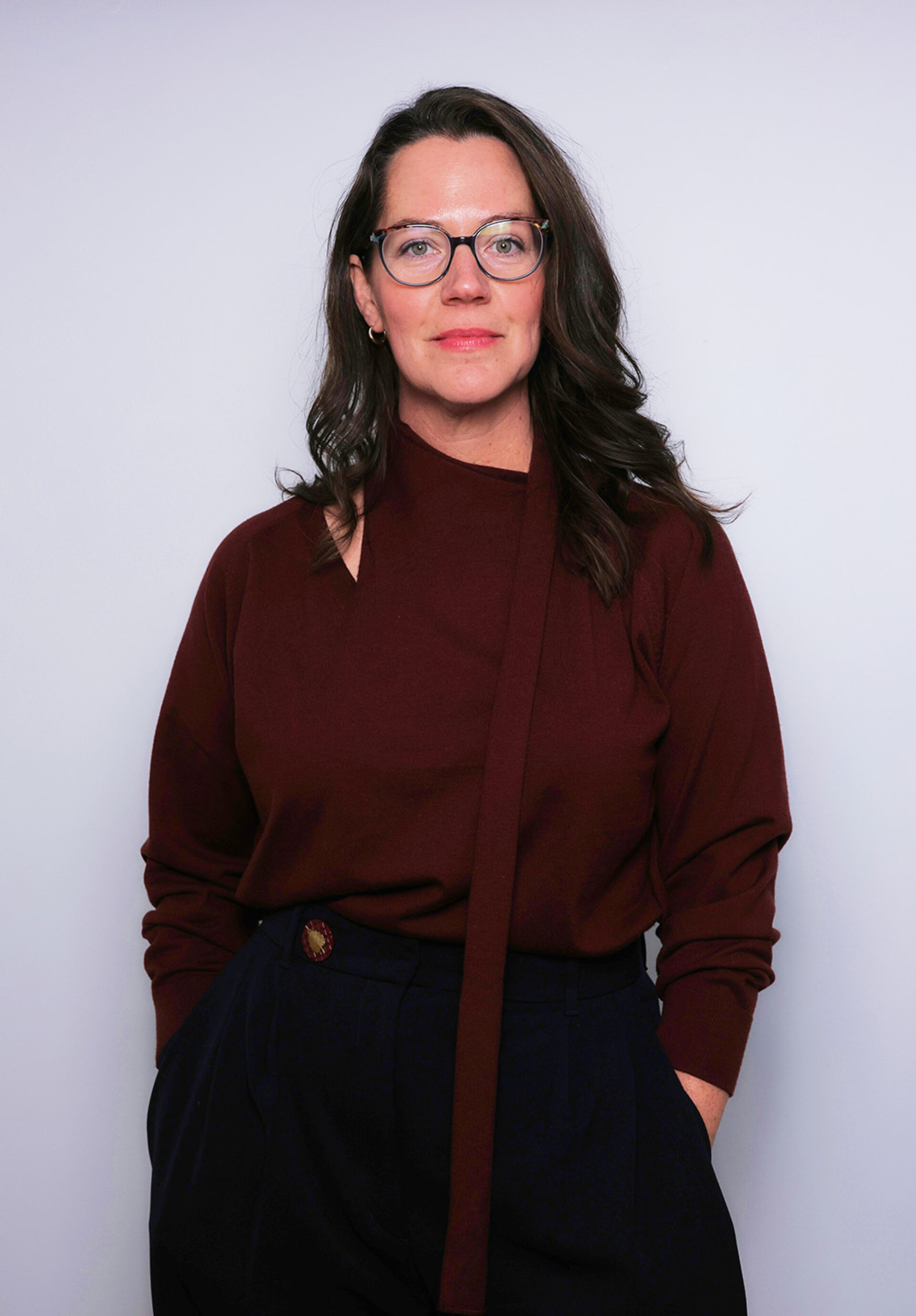 Woman with shoulder-length wavy brown hair and round glasses wearing a burgundy sweater and navy trousers, hands in pockets, neutral smile.
