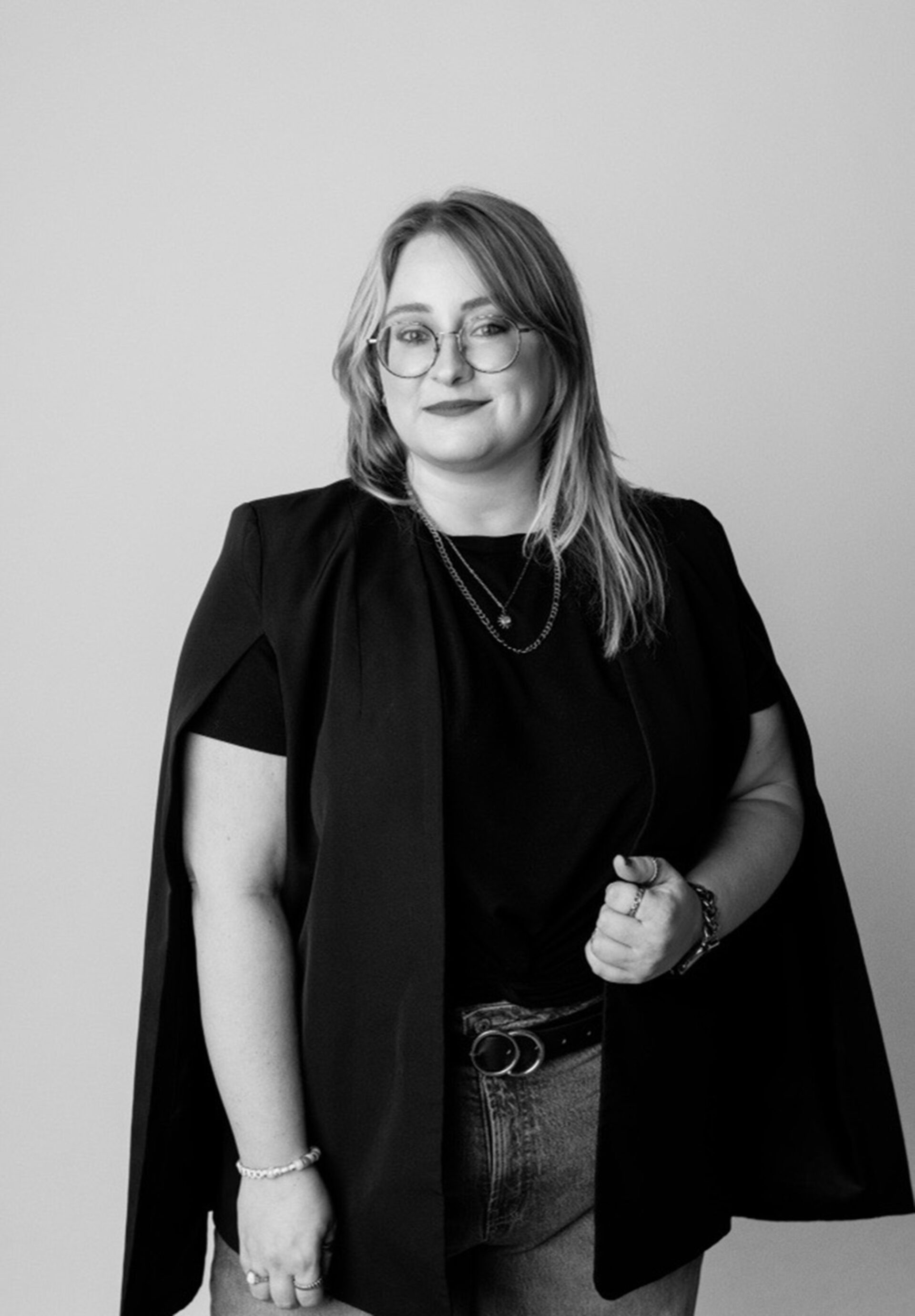 Black-and-white portrait of a smiling woman with round glasses, shoulder-length hair, layered necklaces, cape-style blazer, jeans and rings.