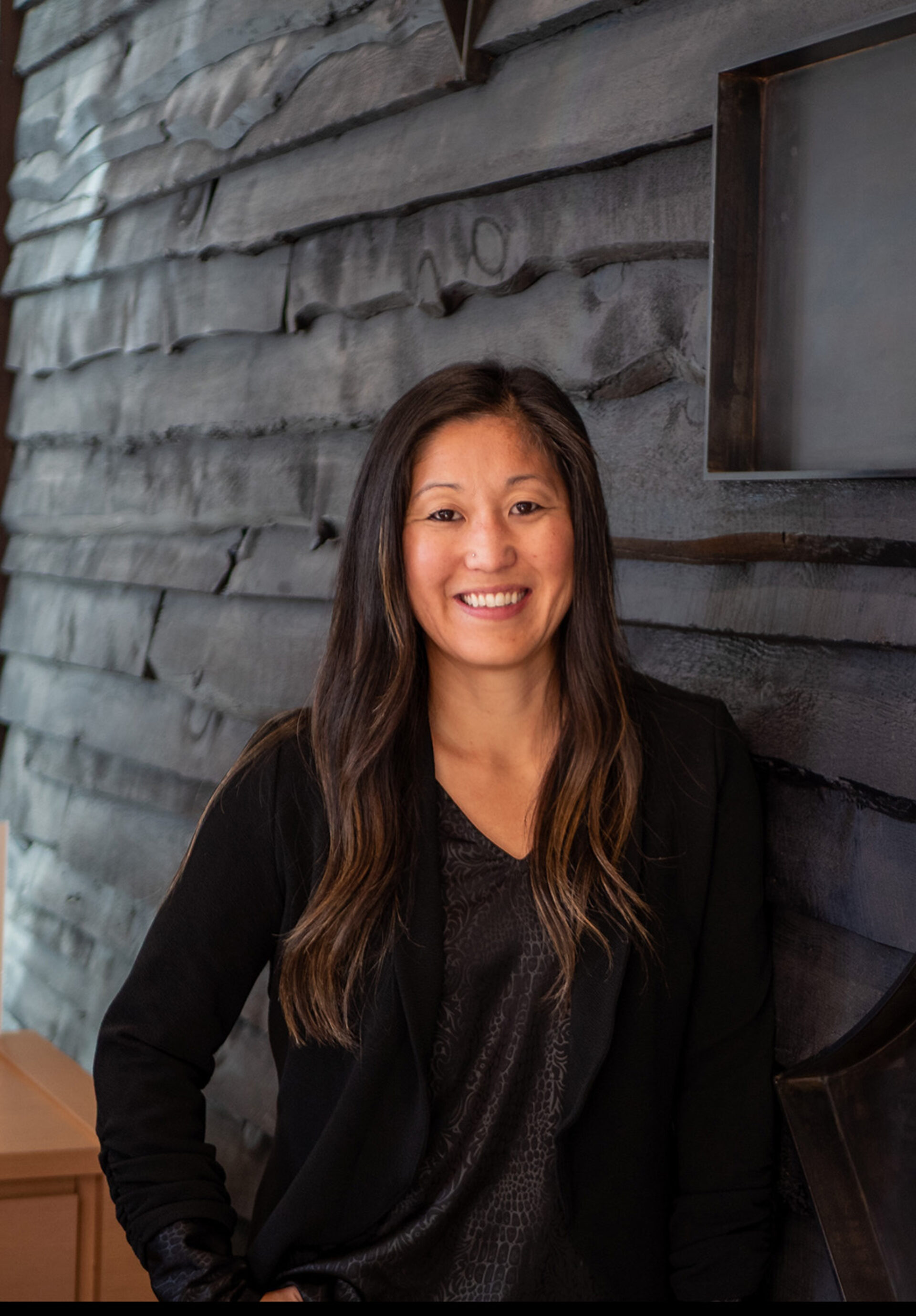 Smiling woman with long dark highlighted hair in a black blazer and patterned blouse, leaning against a textured dark shingle wall.