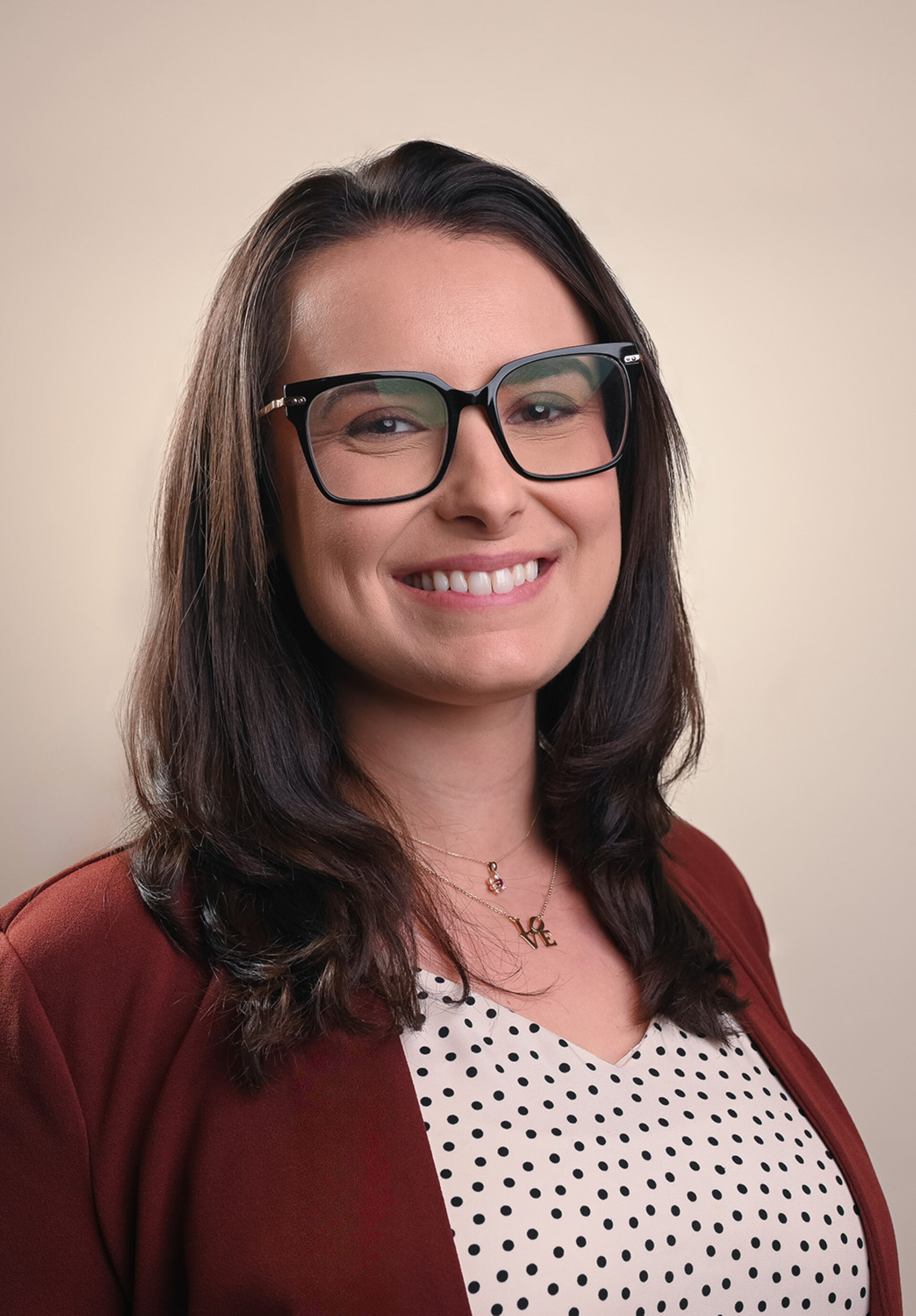 Smiling woman with shoulder-length dark hair and black-framed glasses wearing a burgundy blazer, polka-dot blouse and gold LOVE necklace.