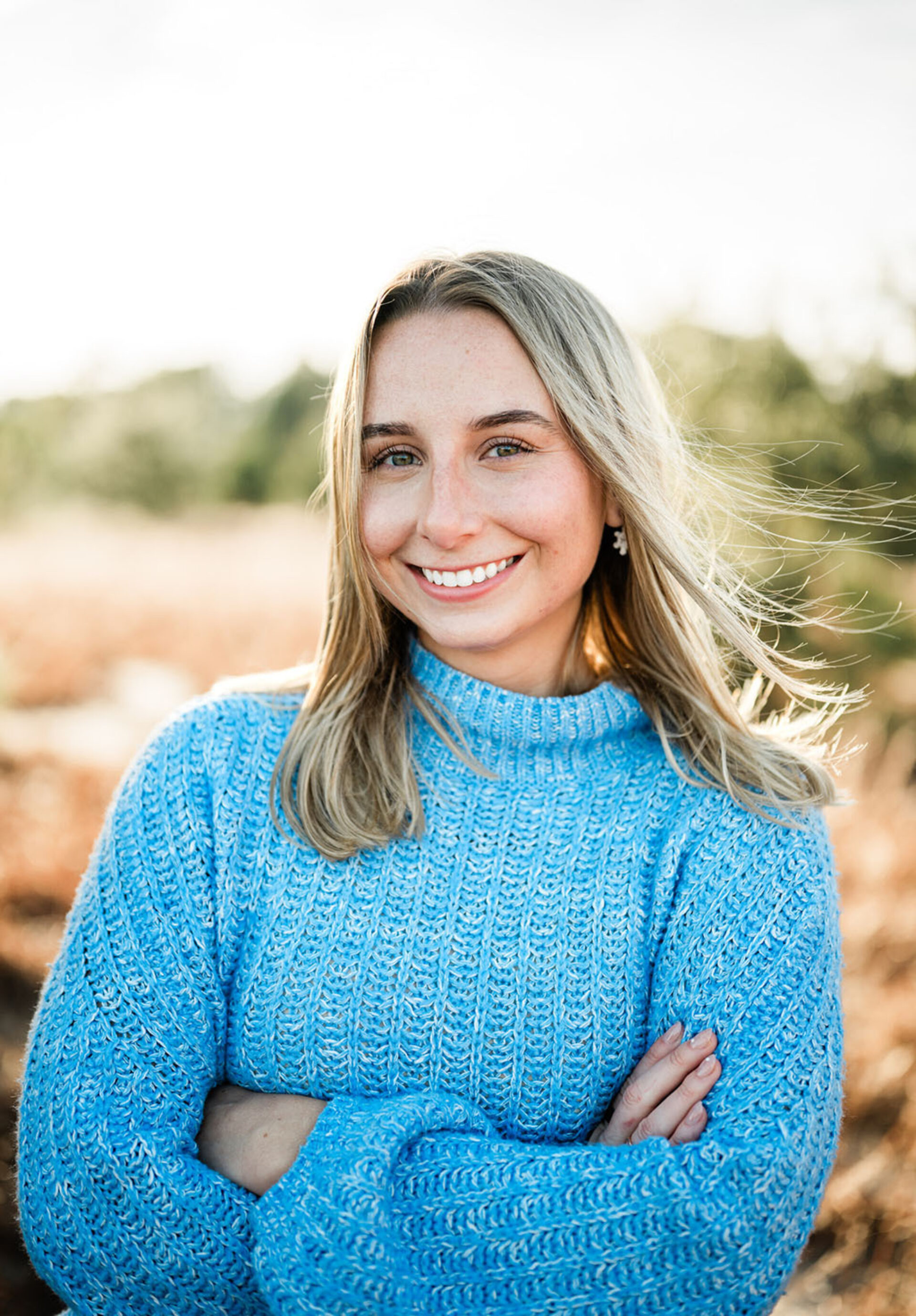 Young blonde woman outdoors smiling with arms crossed, wearing a sky-blue sweater as wind lifts her hair against a sunlit field.