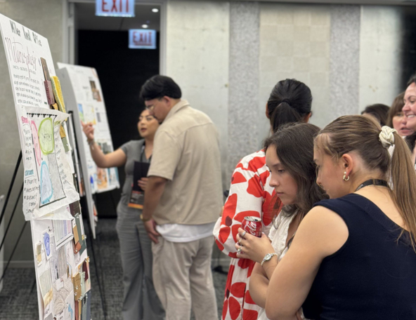 Conference attendees gather at design posters in a meeting room, studying sketches and notes while one woman holds a Coca-Cola can.
