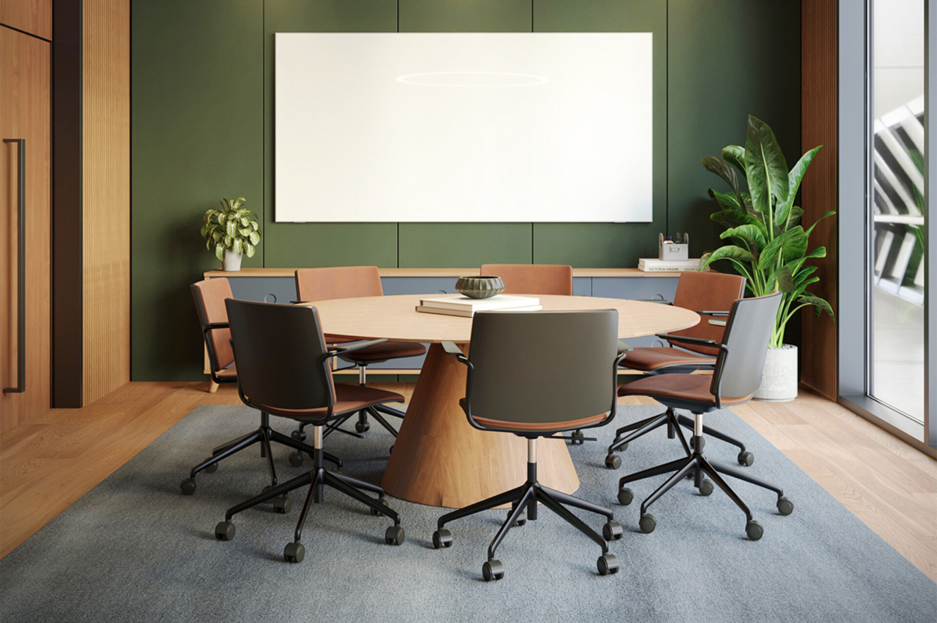 Modern meeting room with round wooden table on cone base, six swivel chairs on rug, olive green paneled wall, blank whiteboard, potted plants and large window.