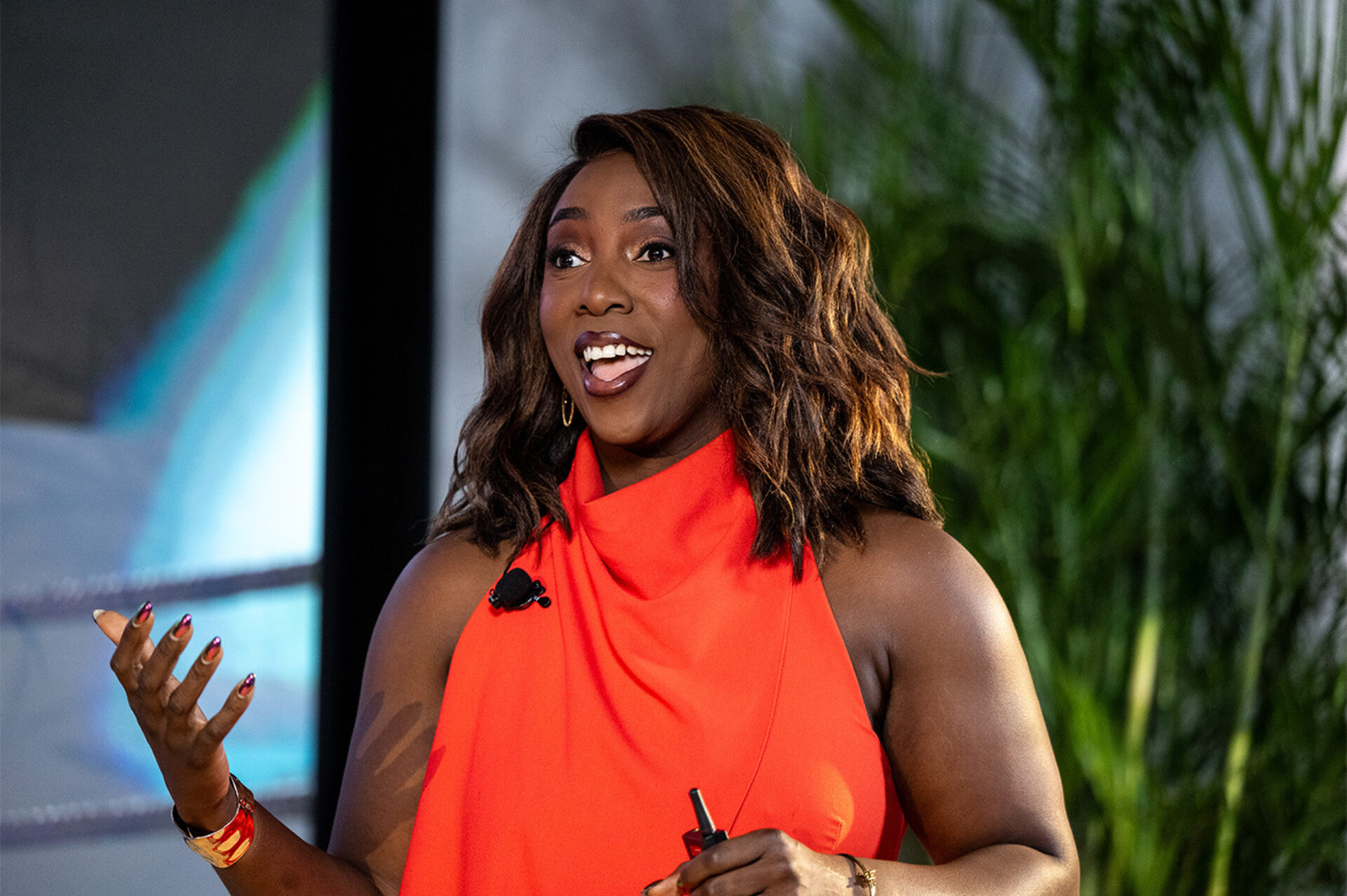 Smiling woman in a sleeveless bright red dress gestures with painted nails while speaking into a lapel mic and holding a clicker.