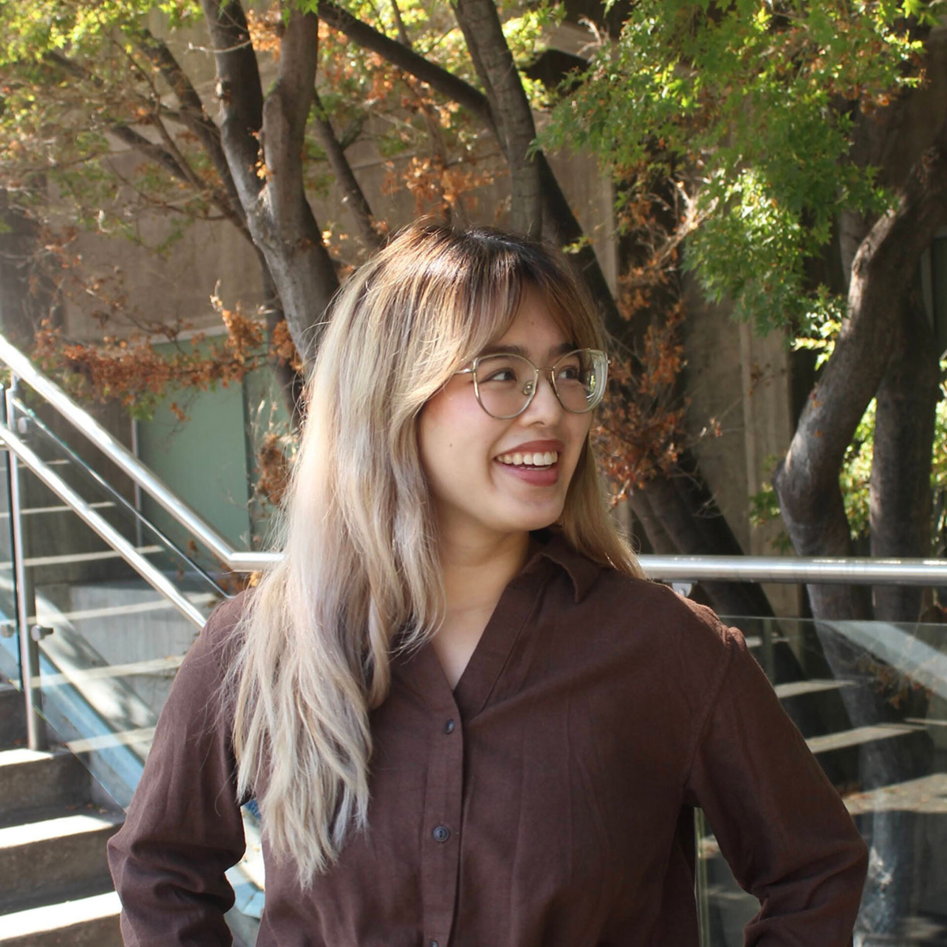 Young woman with long ombre hair and round glasses smiling to the side, wearing a brown button-up, standing by outdoor stairs and trees.