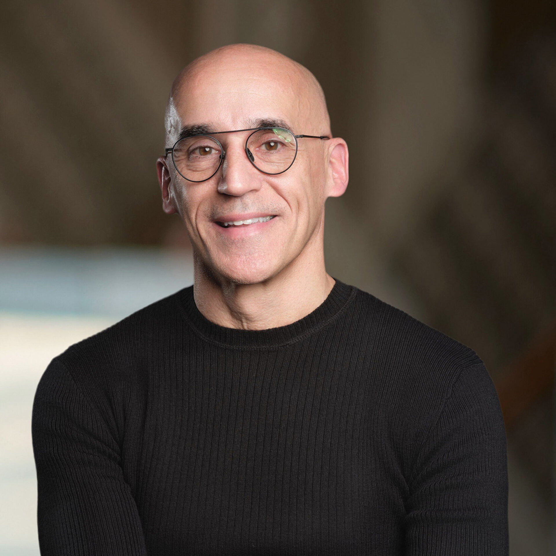 Bald middle-aged man wearing round eyeglasses and a black ribbed sweater, smiling warmly with arms crossed against blurred background.