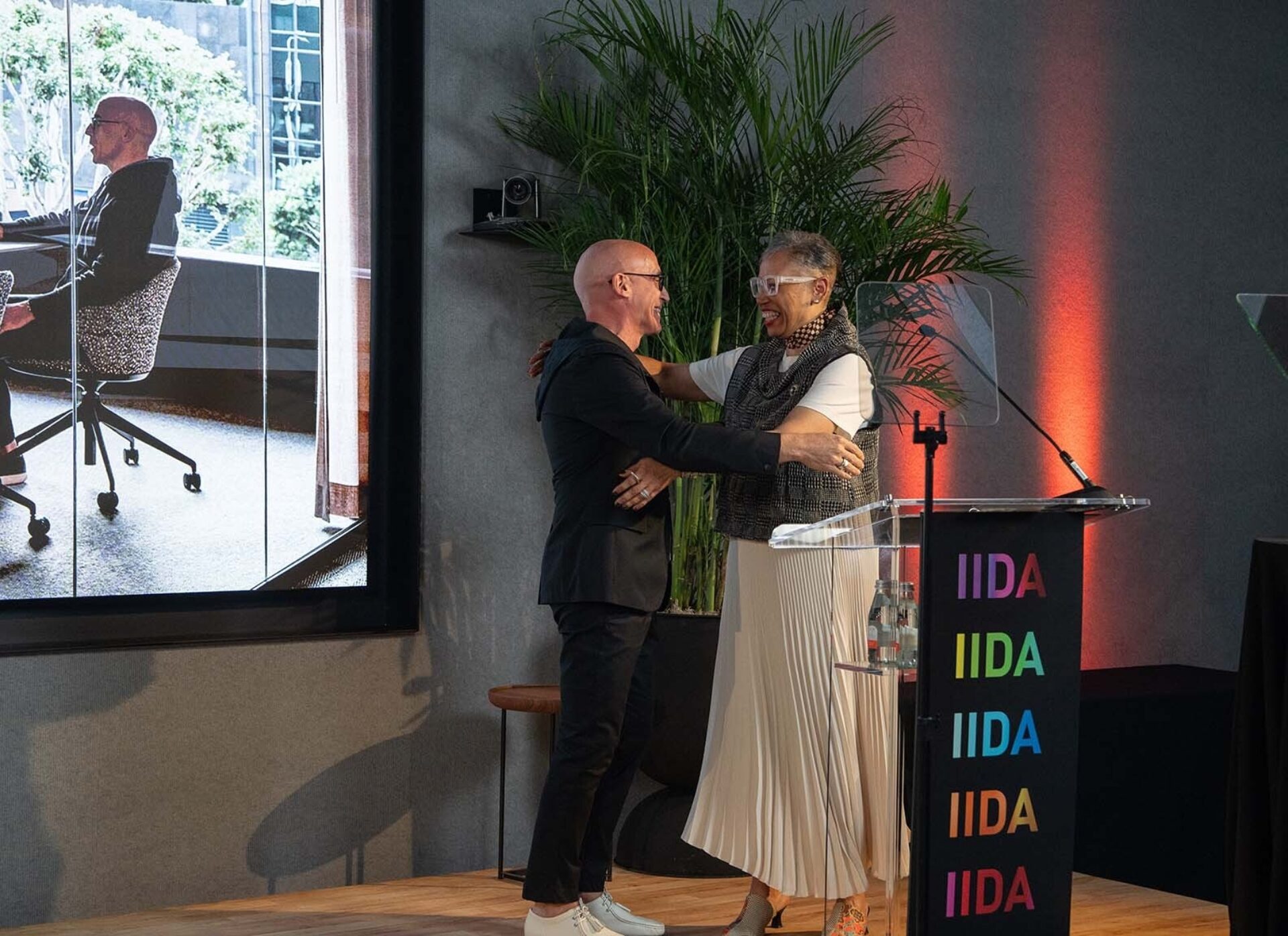 Bald man in black embraces smiling woman with short gray hair, white glasses and pleated skirt beside a podium labeled IIDA onstage.