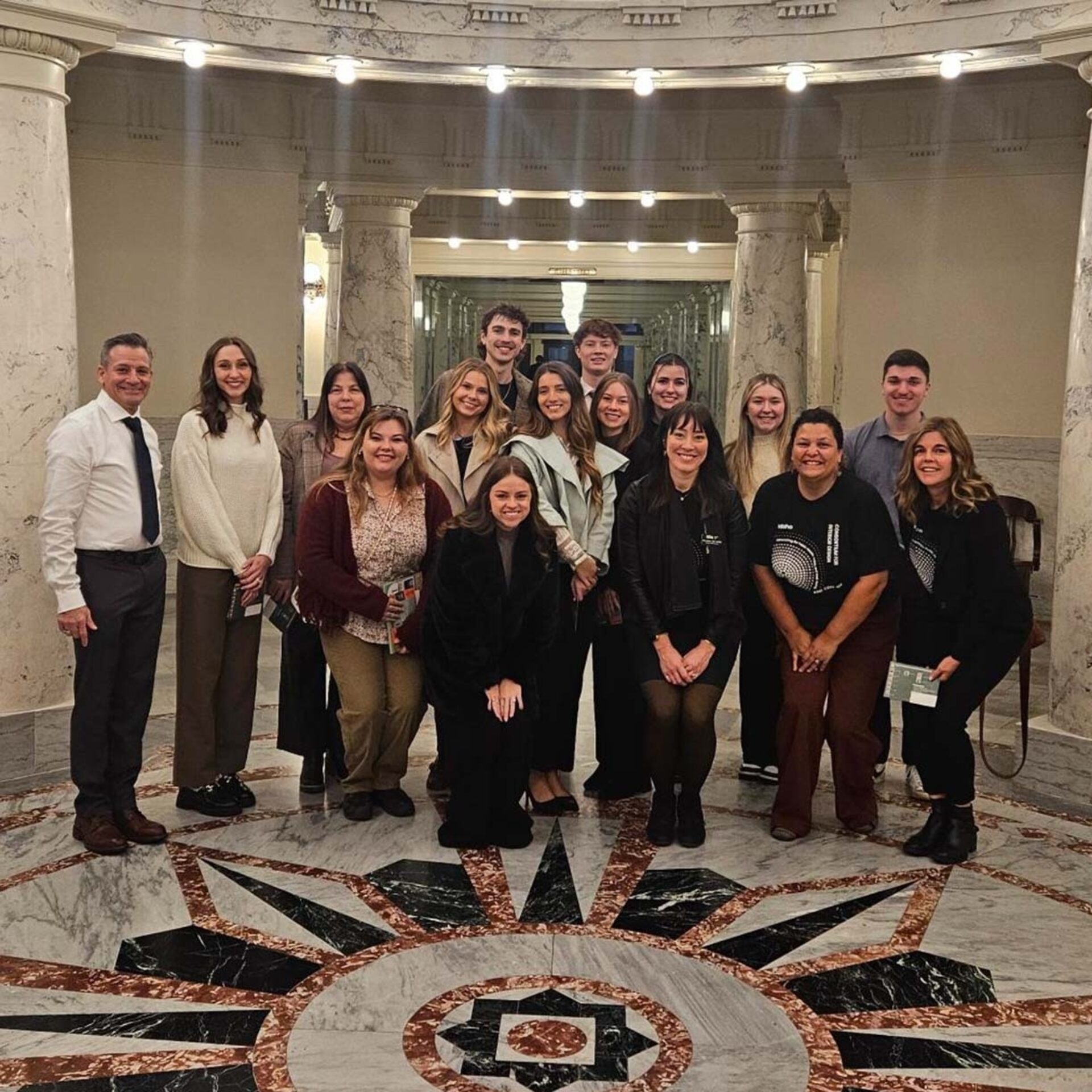 A diverse group of young adults and adults poses together in an ornate marble hall with decorative architecture and lighting.