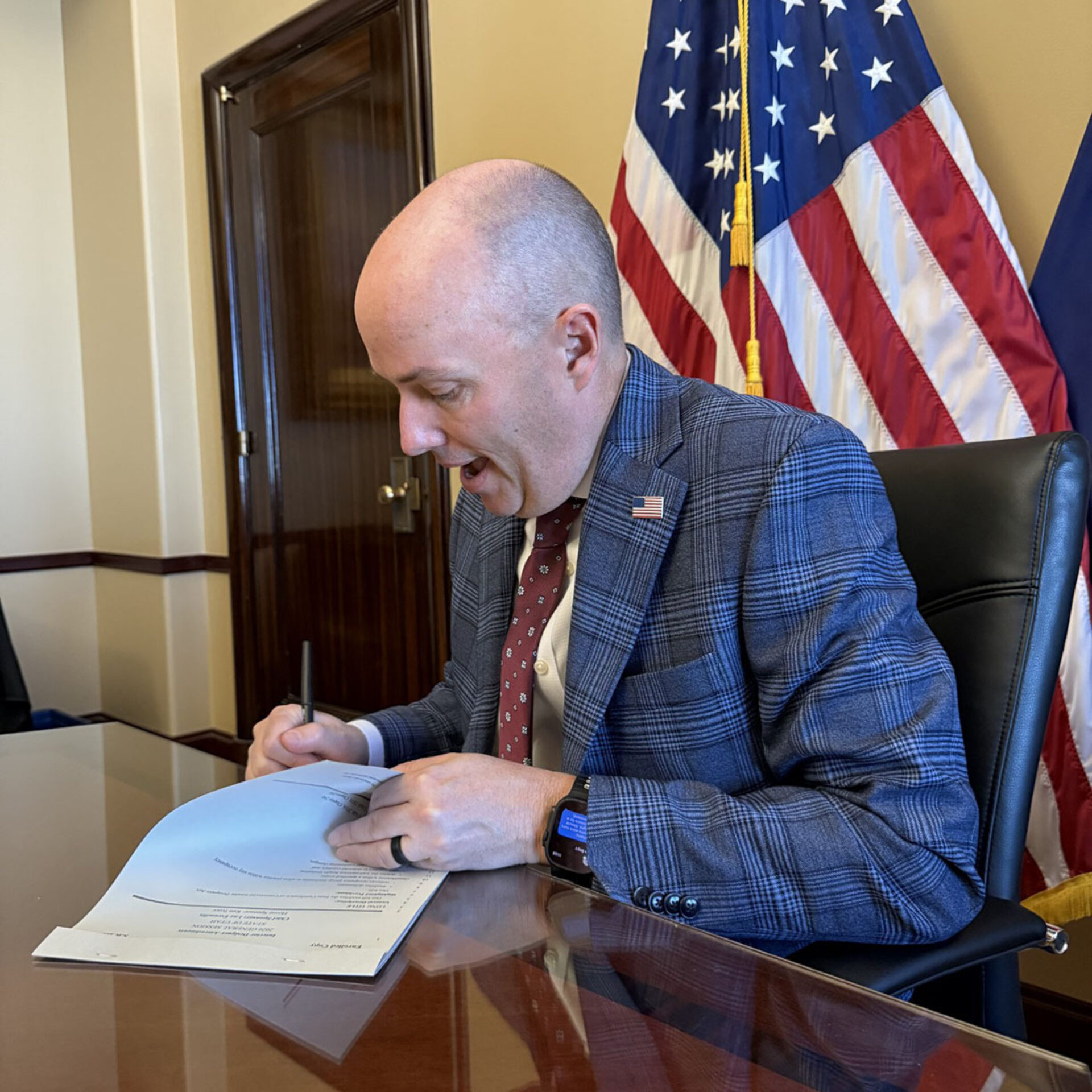 A man in a plaid suit and red tie is signing documents at a desk, with American flags in the background and a focused expression.