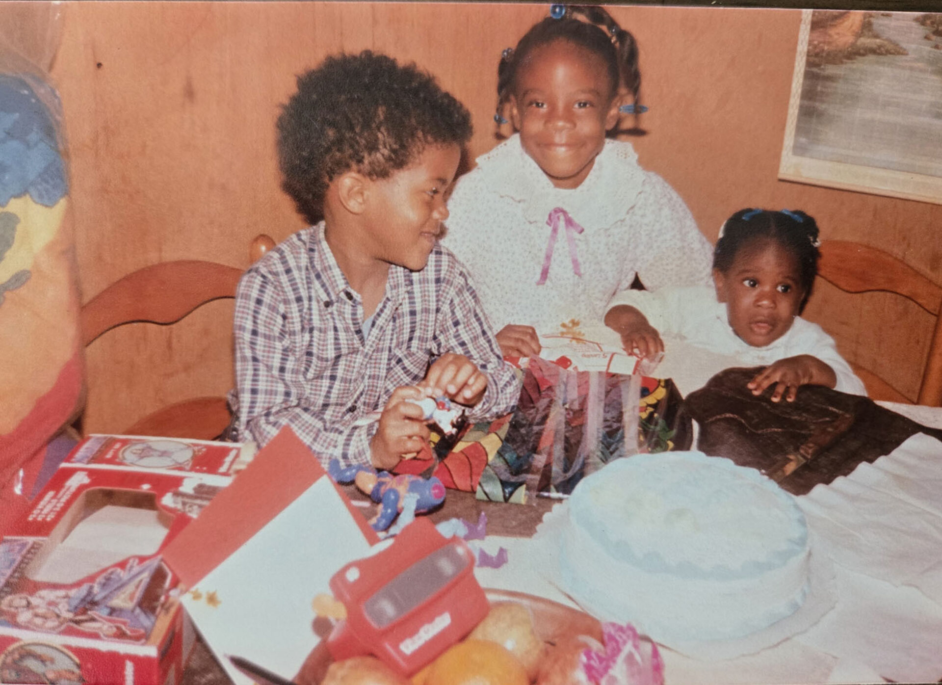 Three children gather around a festive table adorned with a cake, wrapped gifts, and fruit, capturing a joyful celebration moment.