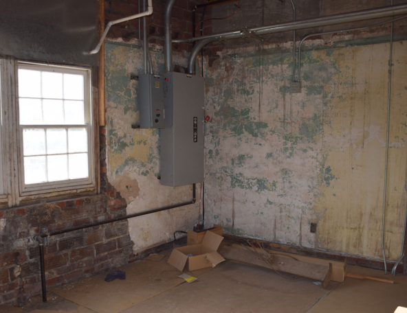 A room with peeling paint, exposed brick, and a gray electrical panel on the wall. Windows allow natural light to enter.