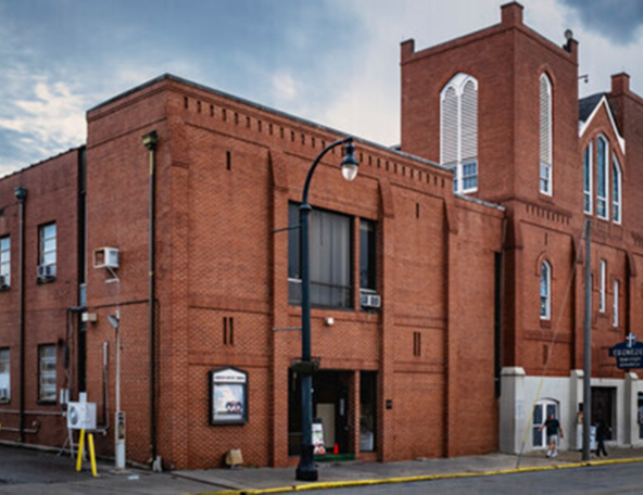 Brick building with large windows and a sign for a church, featuring people walking nearby under a cloudy sky.