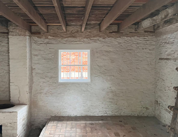 A sparse, rustic room features a stone wall, a wooden ceiling, a small window with a view of brick buildings, and a brick floor.