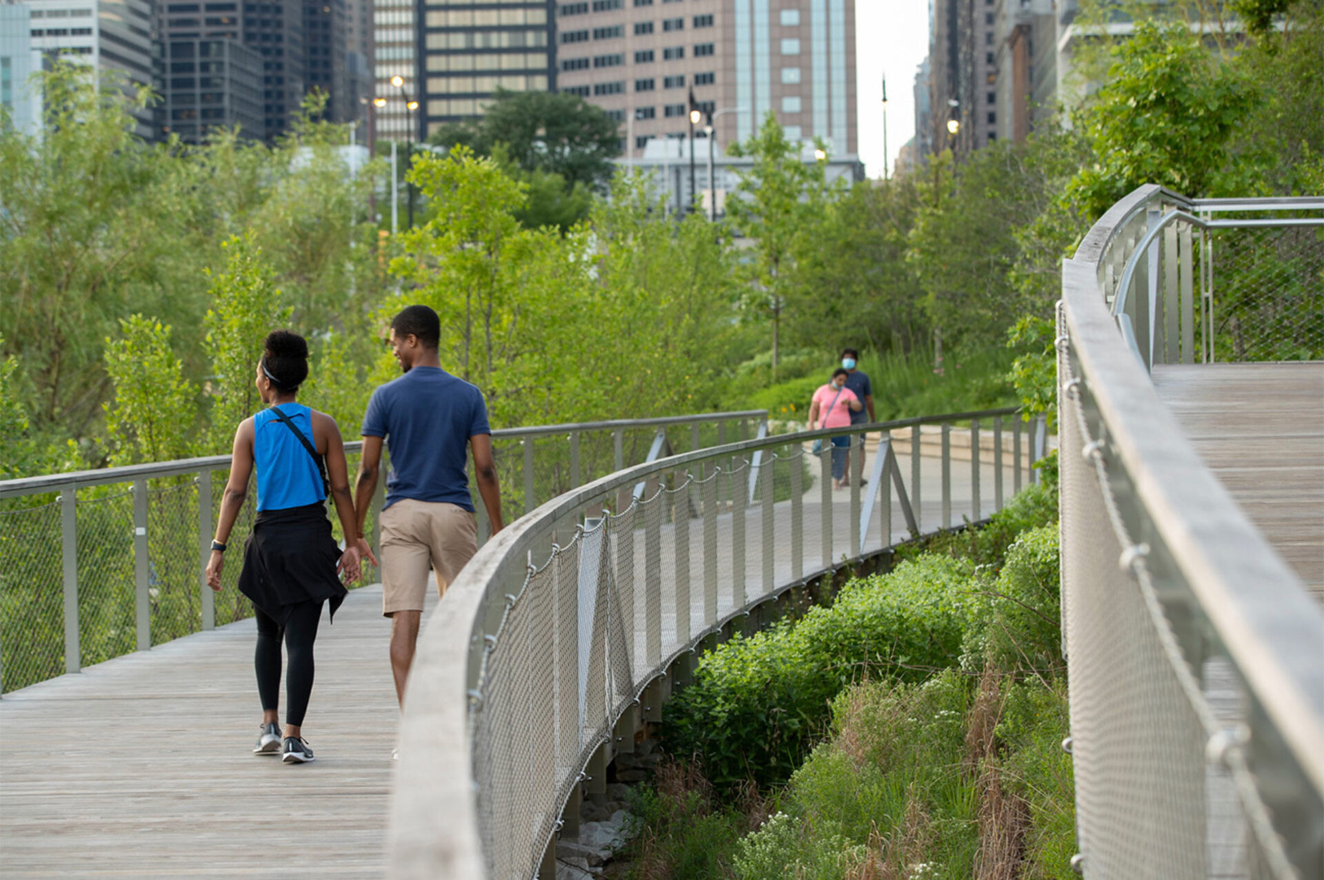 A couple walks hand in hand along a winding path with lush greenery, while another pair strolls in the background among city skyscrapers.