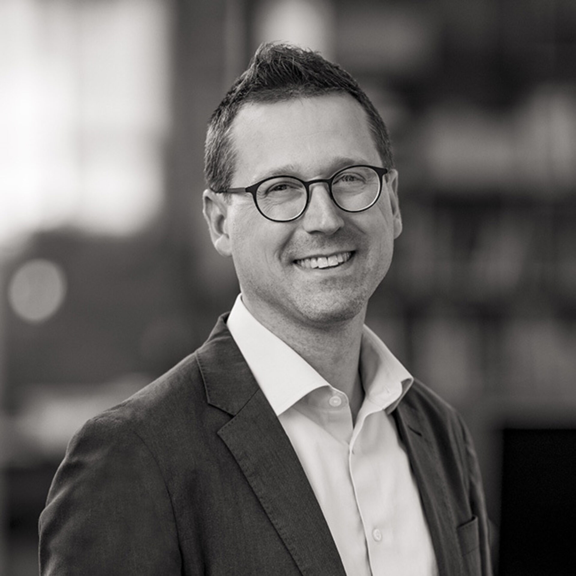 A smiling man with glasses and short hair wears a dark suit over a light shirt, posing in a softly blurred indoor setting.