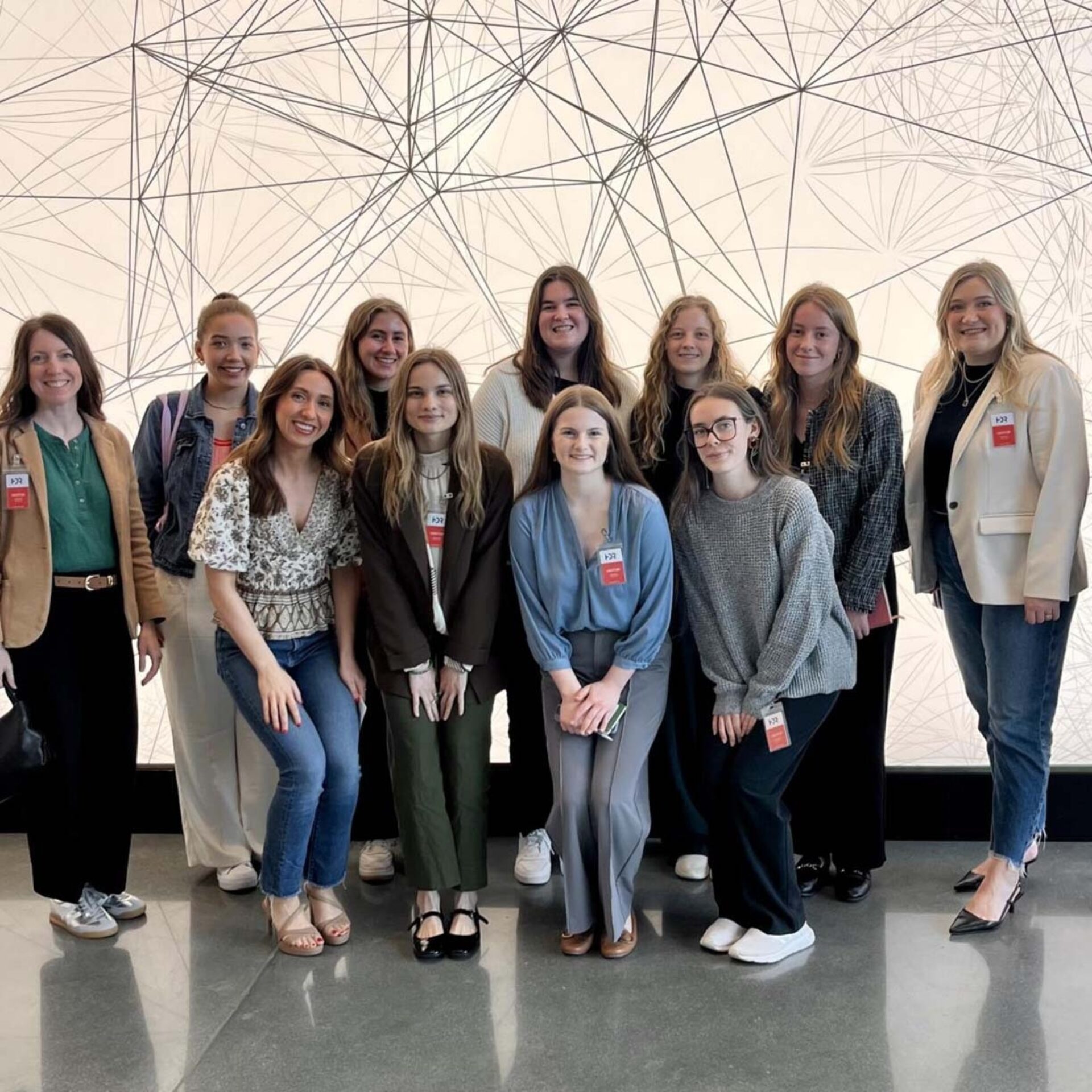 A diverse group of twelve women poses together, wearing name tags and standing in front of an abstract, linear background.