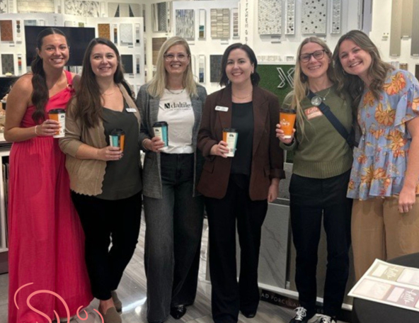 A group of six women stands together in a design showroom, holding cups, with colorful tiles and materials displayed in the background.