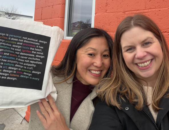 Two women pose for a selfie, holding a tote bag with colorful text about the importance and significance of design against an orange wall.