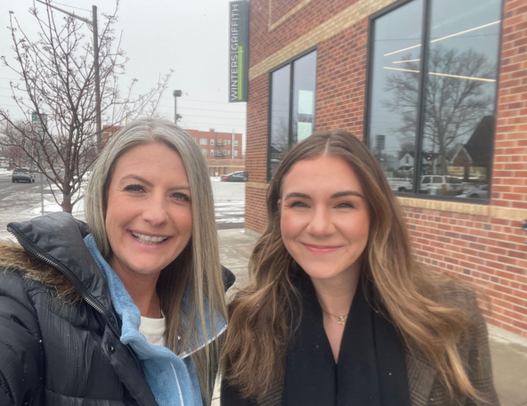 Two women smile together outdoors near a brick building, with a winter environment featuring light snow and bare trees in the background.