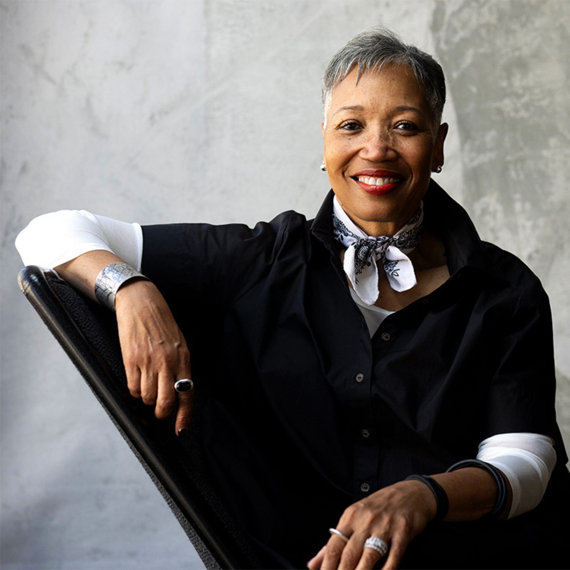 Smiling older woman with short gray hair and red lipstick, wearing a black shirt, white neckerchief and silver jewelry, seated relaxed.